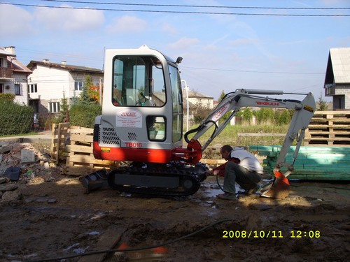 Operator myje małą koparkę Takeuchi TB016 na placu budowy, widoczne palety i gruz w tle.